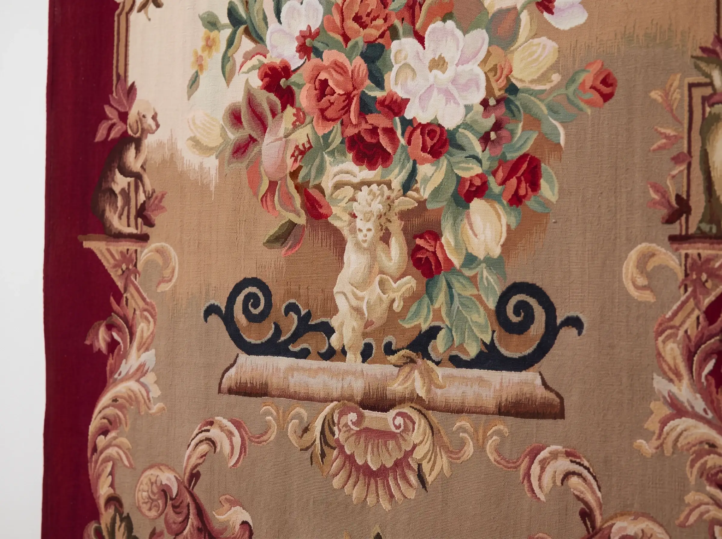 Burgundy Rose Urn Wall Tapestry 5 Close-up of a woven cherub holding an urn of red roses, white blooms and tulips above navy scrollwork on a ledge