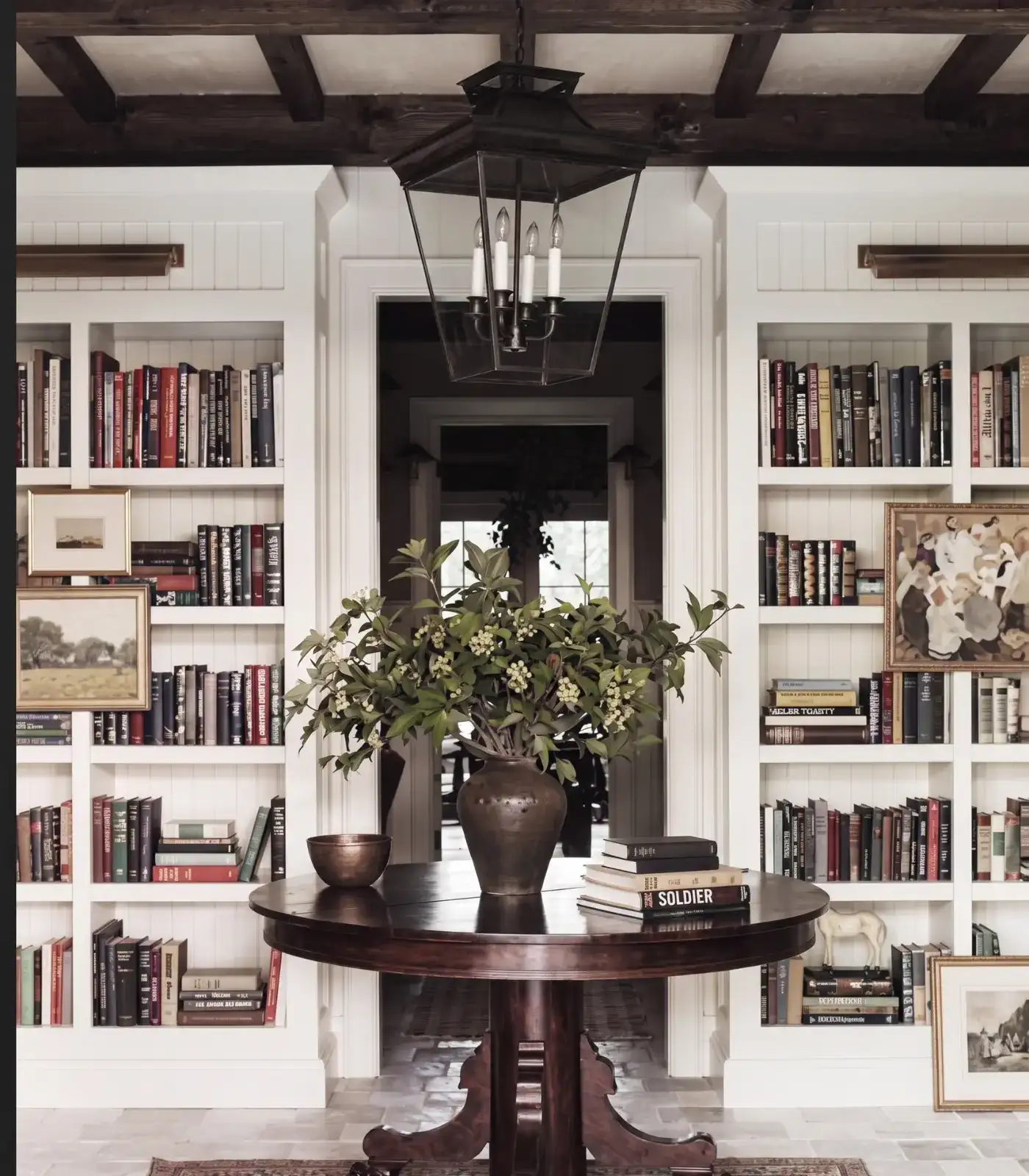 Writer's cottage library with round dark pedestal table, white built-in bookshelves, dark beamed ceiling, iron lantern — balance without matching